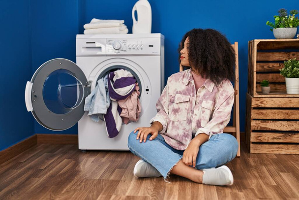 Молода афроамериканська жінка, яка сидить на підлозі і займається пранням, дивлячись убік, відпочиває профіль з природним обличчям з упевненою посмішкою..  - Фото, зображення