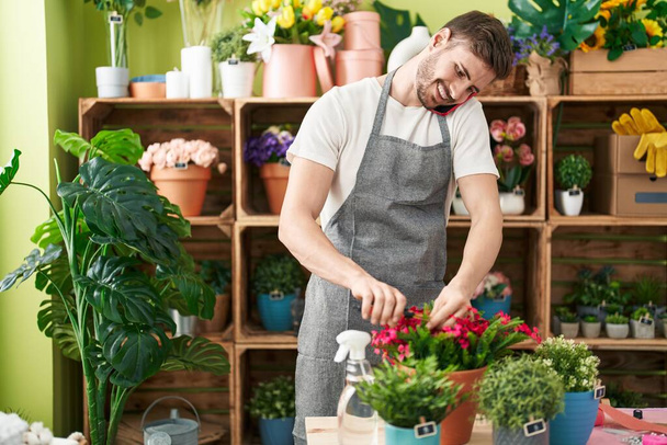 Young caucasian man florist talking on smartphone cutting plant at flower shop - Foto, Bild