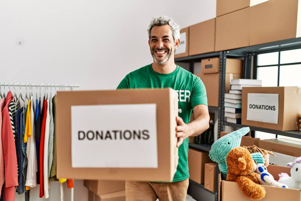 Keski-ikäinen harmaa tukka mies vapaaehtoinen hymyilee luottavainen tilalla lahjoituksia paketti hyväntekeväisyyskeskuksessa - Valokuva, kuva