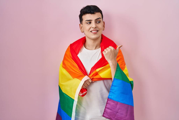 Persona non binaria in possesso di bandiera arcobaleno lgbtq puntando il pollice verso l'alto per il lato sorridente felice con la bocca aperta  - Foto, immagini