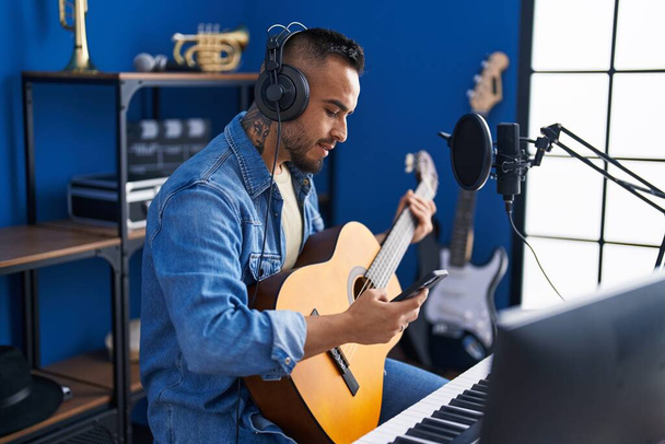 音楽スタジオでクラシックギターを演奏するスマートフォンを使用して若いヒスパニック系の男性アーティスト - 写真・画像