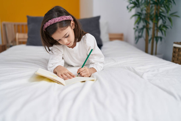 Adorable hispanic girl writing on notebook lying on bed at bedroom - Fotografie, Obrázek