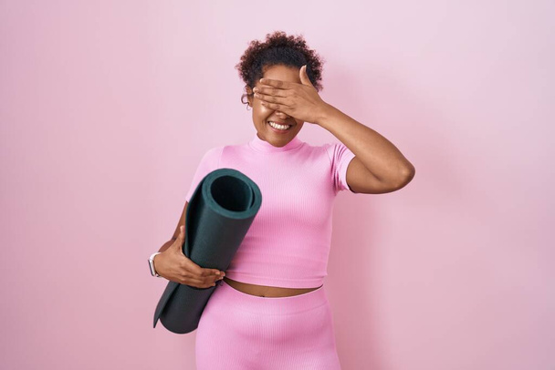 Junge hispanische Frau mit lockigem Haar hält Yogamatte über rosa Hintergrund lächelnd und lachend mit der Hand auf Gesicht bedeckt Augen für Überraschung. Blindes Konzept.  - Foto, Bild