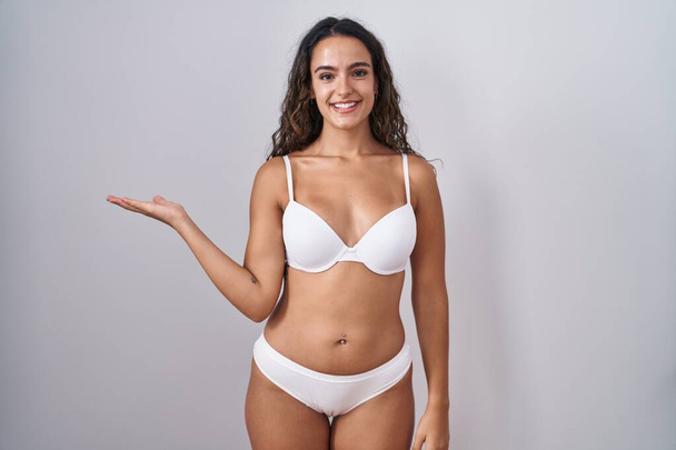 Young hispanic woman wearing white lingerie smiling cheerful presenting and pointing with palm of hand looking at the camera.  - Photo, Image