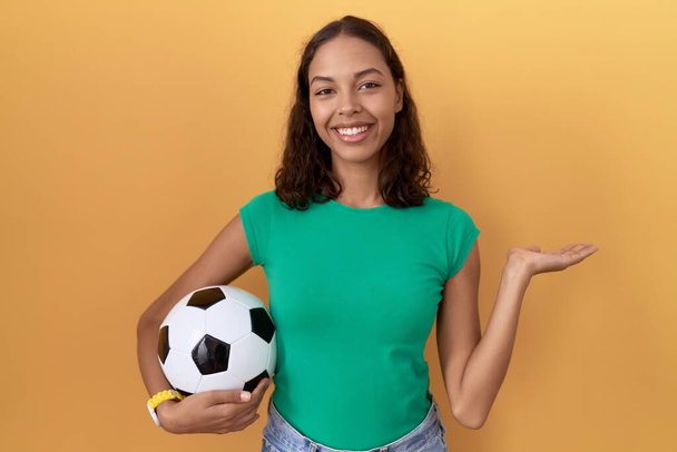 Young hispanic woman holding ball smiling cheerful presenting and pointing with palm of hand looking at the camera.  - Photo, Image