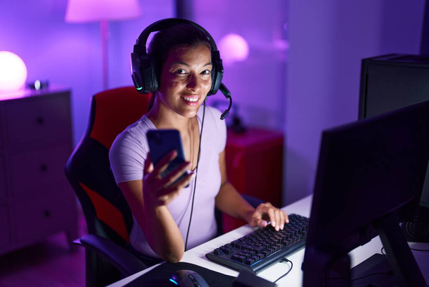 Young beautiful hispanic woman streamer using computer and smartphone at gaming room - Fotoğraf, Görsel