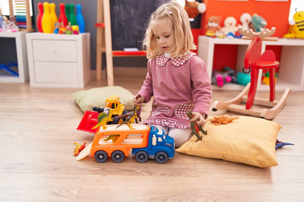 Чарівна блондинка грає з іграшкою вантажівки та динозавра, сидячи на підлозі в дитячому садку
 - Фото, зображення
