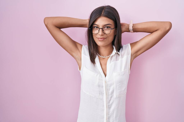 Brunette young woman standing over pink background wearing glasses relaxing and stretching, arms and hands behind head and neck smiling happy  - Фото, зображення