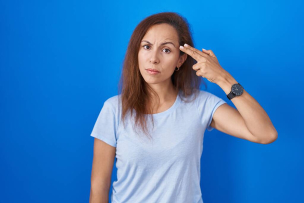 Brunette woman standing over blue background shooting and killing oneself pointing hand and fingers to head like gun, suicide gesture.  - Photo, Image