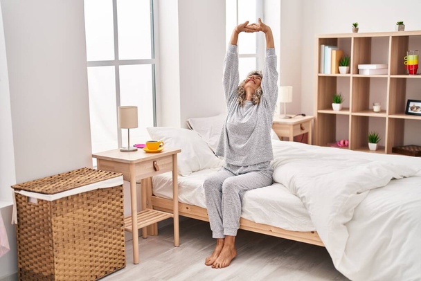 Mulher de meia idade esticando os braços sentados na cama no quarto - Foto, Imagem