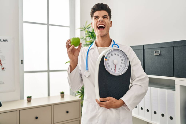Young hispanic doctor man holding scale at dietitian clinic angry and mad screaming frustrated and furious, shouting with anger looking up.  - Photo, Image