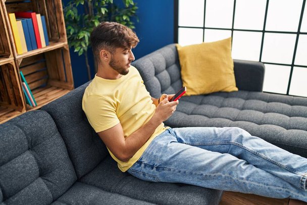 Young arab man using smartphone sitting on sofa at home - Photo, Image
