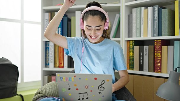 Young beautiful hispanic woman student listening to music dancing at library university - 写真・画像