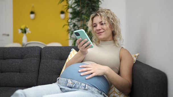 Young pregnant woman using smartphone touching belly at home - Fotoğraf, Görsel