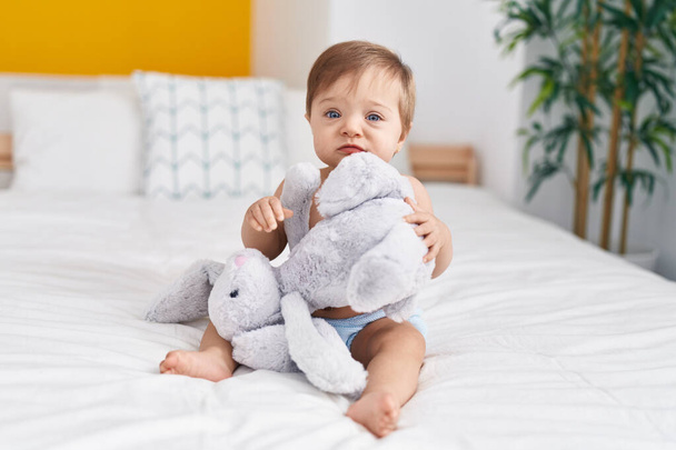 Liebenswertes kaukasisches Baby sitzt auf Bett und spielt mit Puppe im Schlafzimmer - Foto, Bild