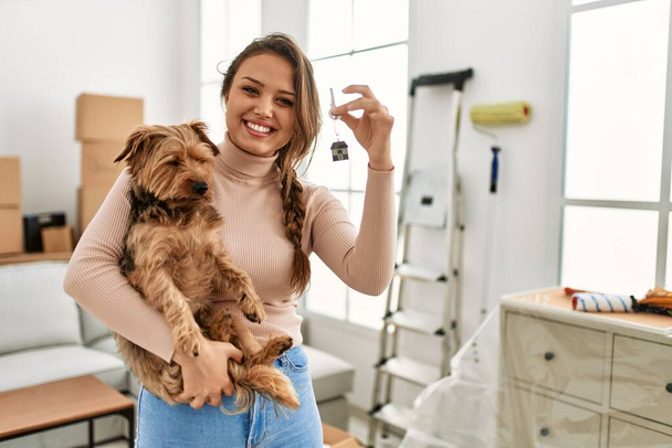 Young beautiful hispanic woman hugging dog holding key at new home - Фото, зображення