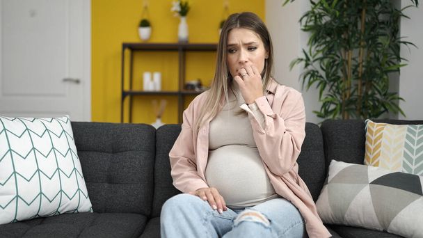 Mujer embarazada joven tocando el vientre con expresión preocupada en casa - Foto, Imagen