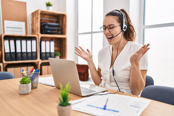 Young hispanic woman wearing call center agent headset at the office celebrating mad and crazy for success with arms raised and closed eyes screaming excited. winner concept  - Photo, Image