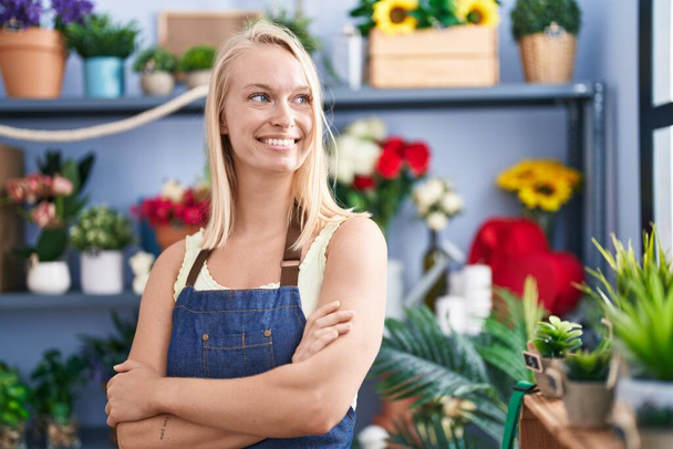 Молода блондинка флорист посміхається впевнено стоячи з обіймами схрестив жест у квітковому магазині
 - Фото, зображення