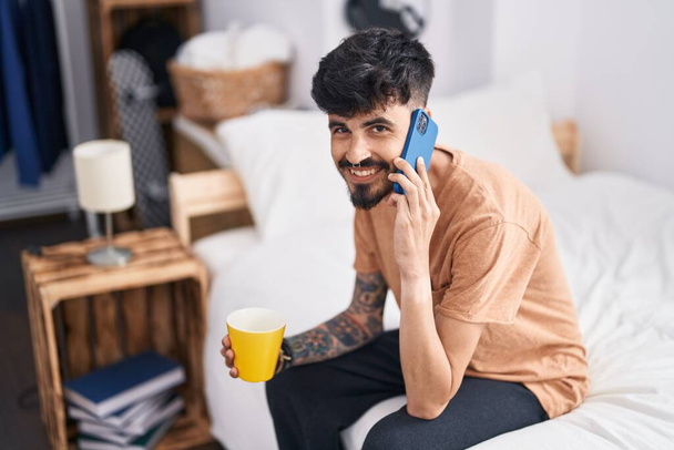 Hombre hispano joven hablando en smartphone tomando café en el dormitorio - Foto, imagen