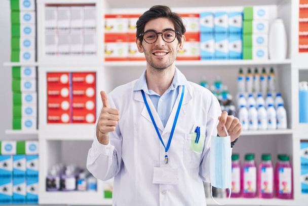 Молодой латиноамериканец, работающий в аптеке, держит защитную маску, улыбается счастливым и позитивным, большой палец вверх делает отличные и одобрения знак  - Фото, изображение