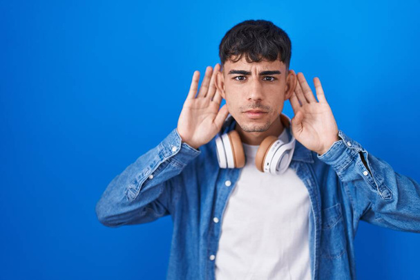 Junger hispanischer Mann, der vor blauem Hintergrund steht und versucht, beide Hände am Ohr zu hören, neugierig auf Klatsch. Hörprobleme, Gehörlosigkeit  - Foto, Bild