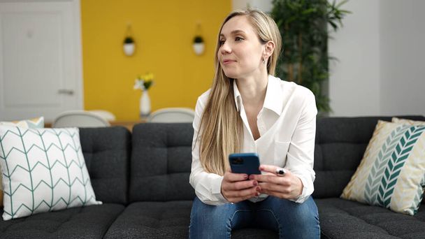 Νεαρή ξανθιά γυναίκα που χρησιμοποιεί το smartphone κάθεται στον καναπέ στο σπίτι - Φωτογραφία, εικόνα