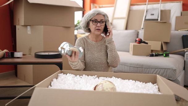 Keski-ikäinen nainen, jolla on harmaat hiukset, puhuu älypuhelimella purkamalla pahvilaatikkoa uudessa kodissa - Valokuva, kuva