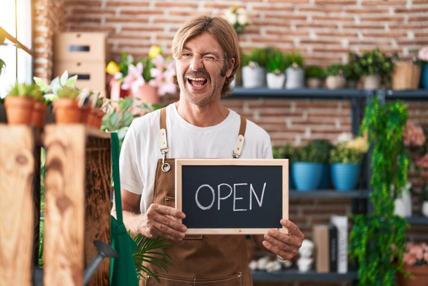 Caucasian man with mustache working at florist holding open sign winking looking at the camera with sexy expression, cheerful and happy face.  - Photo, Image