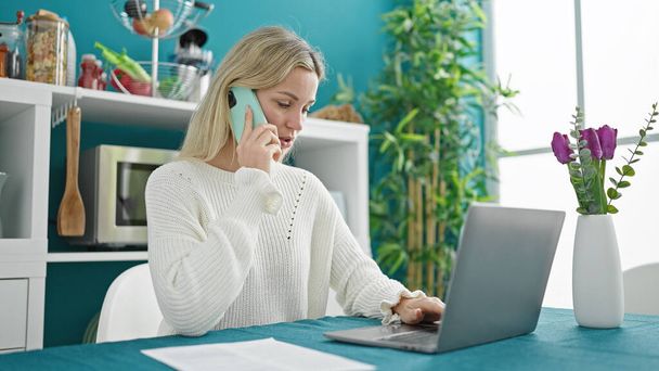 Молодая блондинка с ноутбуком разговаривает на смартфоне в столовой - Фото, изображение