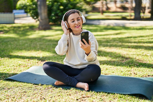 Middle age woman listening to music sitting on herb at park - Photo, image