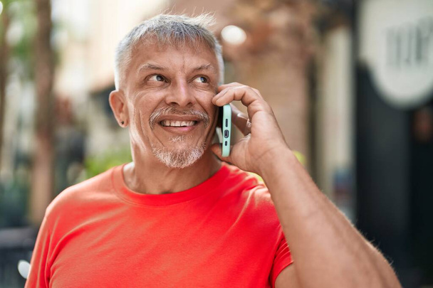 Keski-ikäinen harmaa tukka mies hymyilee luottavainen puhuu älypuhelimeen kadulla - Valokuva, kuva