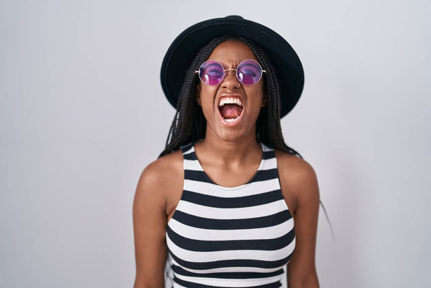 Jovem afro-americana com tranças usando chapéu e óculos de sol com raiva e gritos loucos frustrados e furiosos, gritando com raiva. raiva e conceito agressivo.  - Foto, Imagem