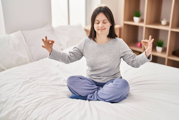 Mujer joven con síndrome de Down haciendo ejercicio de yoga sentada en la cama en el dormitorio - Foto, imagen