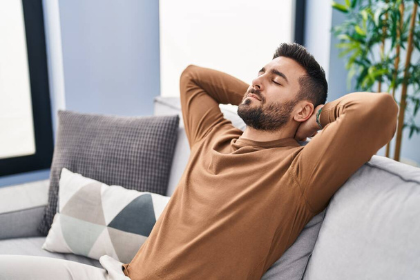Young hispanic man relaxed with hands on head sitting on sofa at home - Photo, Image