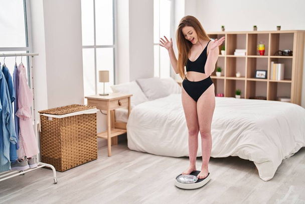 Young caucasian woman standing weight machine to balance weight loss celebrating crazy and amazed for success with open eyes screaming excited.  - Fotografie, Obrázek