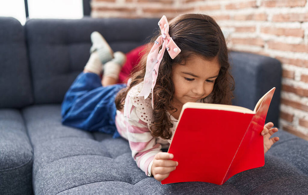 Adorable hispanic girl reading book lying on sofa at home - 写真・画像
