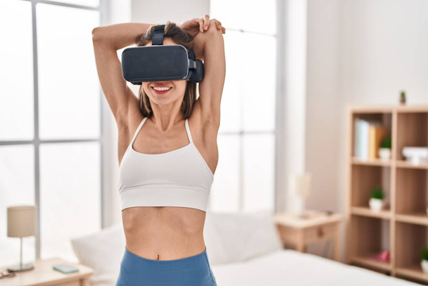 Young beautiful hispanic woman stretching arms using virtual reality glasses at bedroom - 写真・画像