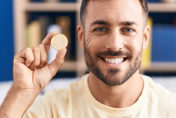 Knappe Spaanse man met tron cryptogeld munt kijken positief en gelukkig staan en glimlachen met een zelfverzekerde glimlach tonen tanden  - Foto, afbeelding