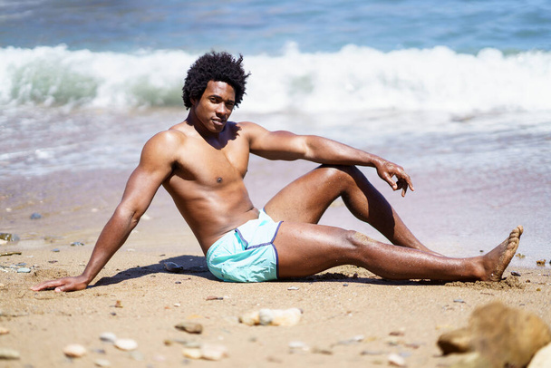 Full body side view van Afro-Amerikaanse man ontspannen op zandstrand met arm op knie terwijl u naar de camera kijkt, en leunend op de hand in de buurt van schuimige zee - Foto, afbeelding