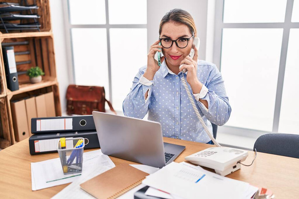 Młoda latynoska bizneswoman pracująca w biurze rozmawiająca przez dwa telefony zrelaksowana z poważnym wyrazem twarzy. proste i naturalne patrzenie w kamerę.  - Zdjęcie, obraz