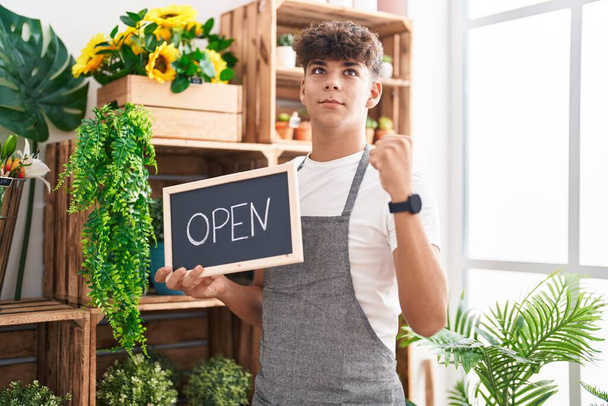 Hispanic teenager working at florist holding open sign screaming proud, celebrating victory and success very excited with raised arms  - Photo, Image
