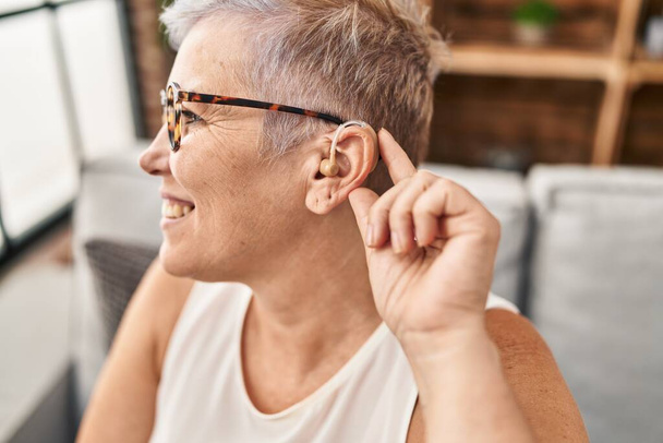 Middle age woman using hearing aid sitting on sofa at home - Foto, Imagem
