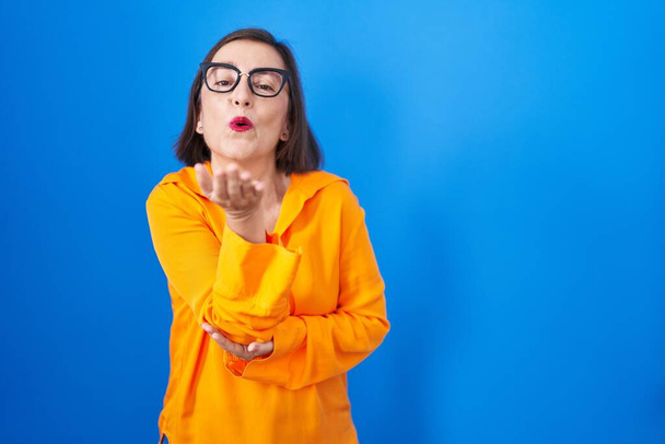 Moyen Age femme hispanique portant des lunettes debout sur fond bleu regardant la caméra souffler un baiser avec la main sur l'air étant belle et sexy. expression d'amour.  - Photo, image