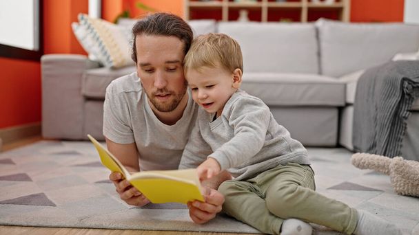 Ojciec i syn czytający książkę, siedzący razem na podłodze w domu. - Zdjęcie, obraz