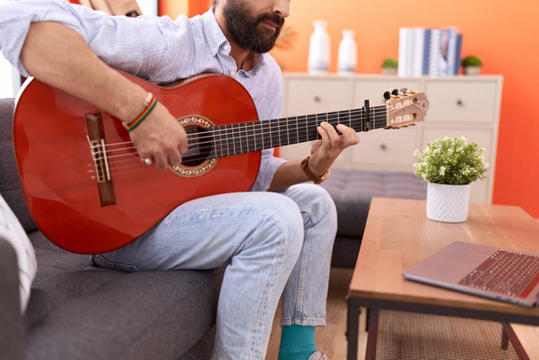Young hispanic man having online guitar class sitting on sofa at home - Foto, afbeelding