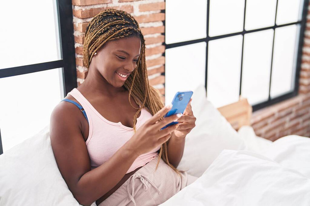 African american woman using smartphone sitting on bed at bedroom - 写真・画像