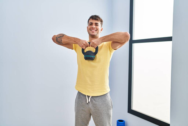 Jeune homme hispanique souriant confiant en utilisant la formation kettlebell au centre sportif - Photo, image