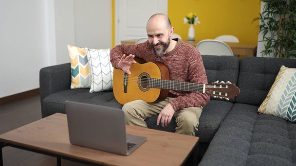 młody łysy człowiek mając online klasyczne lekcje gry na gitarze siedzi na kanapie w domu - Zdjęcie, obraz