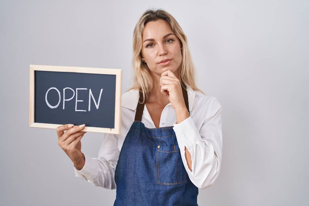 Young blonde woman wearing apron holding blackboard with open word serious face thinking about question with hand on chin, thoughtful about confusing idea  - Foto, Imagem